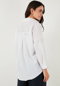 Camicia bianca con bottoni, maniche lunghe, orlo curvo e dettaglio della piega posteriore. Realizzata in tessuto leggero, presenta polsini con bottoni e un colletto classico.