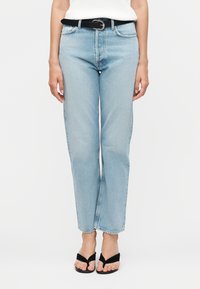 Mujer con vaqueros rectos de pierna en azul claro, cinturón negro, top blanco y sandalias negras con tacón y tiras en los dedos, de pie sobre un fondo blanco.