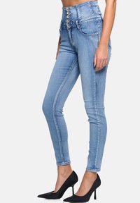 Jeans de tiro alto en denim azul con cierre de cinco botones, corte slim, costuras laterales y una textura suave. Combinados con zapatos de tacón alto negros.