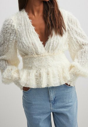 Femme portant un chemisier blanc texturé à manches longues avec un décolleté en V profond et un ourlet à volants, associé à un jean en denim bleu.