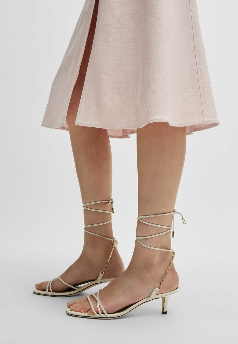 Beige sandaalit, joissa on alhainen korko, nilkkaketjut ja kultaiset yksityiskohdat. Kenkien muotoilu on minimalistinen ja avokärkinen.