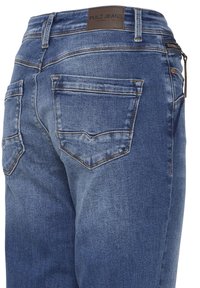 Jean en denim bleu à la texture délavée, poches arrière et une étiquette en cuir marron sur la ceinture portant l'inscription "PULZ JEANS".