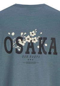 T-shirt di un blu scuro con del bold nero che scrive "OSAKA" e disegni floreali bianchi. Testo aggiuntivo sotto in un font più piccolo. Tessuto morbido.