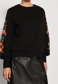 Černá mikina s živým květinovým vyšíváním v oranžové a zelené barvě na rukávech, s pohodlným střihem a kulatým výstřihem.