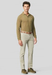 Olijfgroen polo met lange mouwen, lichtbeige broek, bruine riem, donkergroene sneakers; de outfit heeft een subtiele logo op de borst.