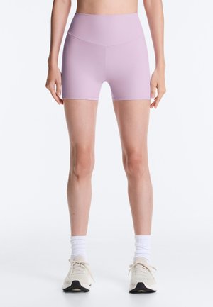 Kobieta ubrana w fioletowe, wysokie szorty rowerowe, białe skarpetki sportowe i beżowe tenisówki, stojąca na tle gładkiej białej ściany.