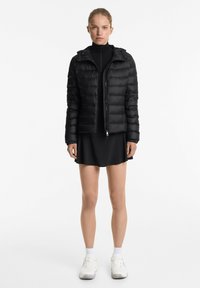 Zwarte pufferjas met capuchon, gequilte ontwerp, volledige rits, elastische manchetten, gedragen over een zwarte jurk, gecombineerd met witte sneakers.