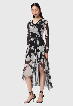 Kobieta ubrana w czarno-białą, długorękawową sukienkę w kwiaty z asymetrycznym dołem i czarne sandały na wysokim obcasie na jednolitym tle.