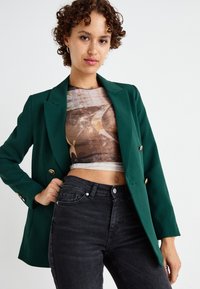Zelený blejzr s dvojitým zapínáním, zlatými knoflíky a klopovými límci. Nosený přes vzorovaný crop top, v kombinaci s černými džíny s vysokým pasem.