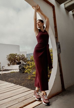 Bordeauxkleurige mouwloze jurk met een gestrikt middel; gedragen met bruin-witte sneakers. Heeft een cirkelvormig architecturaal element en groene planten.