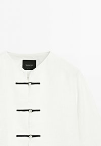 Bílá košile s mandarínským límečkem, čtyřmi vodorovnými černými zapínacími knoflíky vpředu, štítek Massimo Dutti viditelný uvnitř límce.