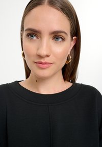 Nahaufnahme einer Person, die ein schwarzes Kleidungsstück mit runder Ausschnitt trägt, das aus glattem Stoff besteht und subtile Nähdetails aufweist. Sichtbare goldene Ohringe in Form eines Rings.