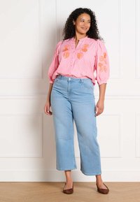Mujer con cabello rizado que lleva una blusa rosa con bordado floral naranja, jeans de pierna ancha azul claro y zapatos planos marrones, de pie en el interior.