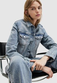 Giacca in denim azzurro chiaro con chiusura a bottoni, due tasche sul petto e texture sbiadita, indossata sopra pantaloni in denim a vestibilità larga.