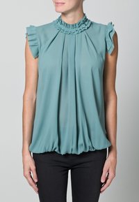Top senza maniche di colore turchese con scollatura arricciata e parte anteriore plissettata, caratterizzato da una vestibilità ampia e orlo arricciato. Indossato con pantaloni scuri.