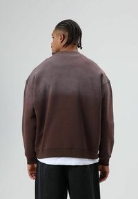 Bruine sweatvest met verloopkleur en raglanmouwen, voorzien van getextureerde accenten en geribbelde manchetten, gecombineerd met losse zwarte denimbroek.