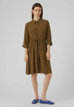 Robe marron à boutons avec col, deux poches poitrine et ceinture à la taille. Tissu fluide avec des manches 3/4. Portée avec des chaussures plates bleues.