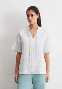 Weißes Leinenoberteil mit V-Ausschnitt und kurzen Ärmeln, ausgestattet mit einer Frontleiste und schmalen Bändern. Getragen mit hellblauen Hosen.