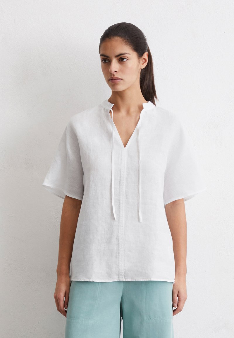 Weißes Leinenoberteil mit V-Ausschnitt und kurzen Ärmeln, ausgestattet mit einer Frontleiste und schmalen Bändern. Getragen mit hellblauen Hosen.