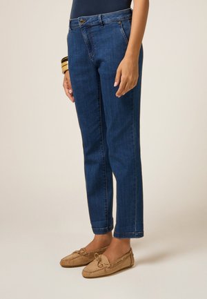 Jeans de mezclilla azul oscuro con corte recto, diseño de cinco bolsillos y cierre de cremallera. Combinados con zapatos estilo mocasín de color marrón claro.