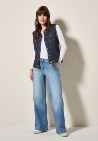 Marinblå denimväst med vit sömnad, framfickor och silverfärgade knappar, kombinerad med ljusblå vida jeans och beige tygskor.
