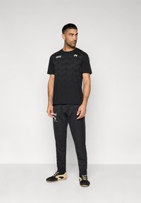 Μαύρο αθλητικό σύνολο που περιλαμβάνει υφασμένη κοντομάνικη μπλούζα και στενά παντελόνια, και τα δύο με λογότυπα. Τα παπούτσια είναι μαύρα και καφέ με κορδόνια.