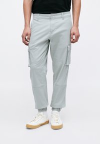 Man som bär ljusgrå cargobyxor med knapp- och dragkedjestängning, elastiska muddar och vita sneakers med gummisulor.