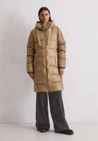 Bēšā pūkainā jaka ar kapuci, ar rāvējslēdzēju priekšā, elastīgām piedurknēm un divām priekšējām kabatām. Nēsāta virs pelēkam platām biksēm un tumšām apaviem.