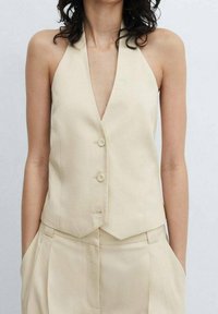 Gilet beige a collo halter con silhouette aderente, caratterizzato da due bottoni e una texture di tessuto liscia, abbinato a pantaloni coordinati.
