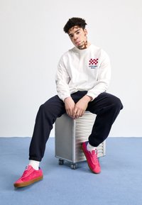 Chemise blanche à manches longues avec motif à carreaux rouge; pantalon foncé; baskets roses avec accents bleus; assis sur une boîte de rangement argentée.