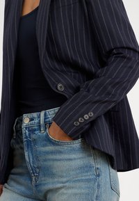 Asmuo dėvi tamsų dryžuotą švarką ant juodo viršaus, ranka įkišta į šviesiai mėlynų džinsų priekinę kišenę.