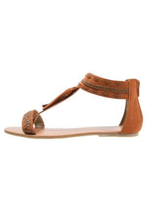 Brązowy zamszowy sandał na płaskiej podeszwie, w stylu T-strap, z plecionym wykończeniem i ozdobami z chwostami. Otwarty nosek i pasek wokół kostki z wycięciami.