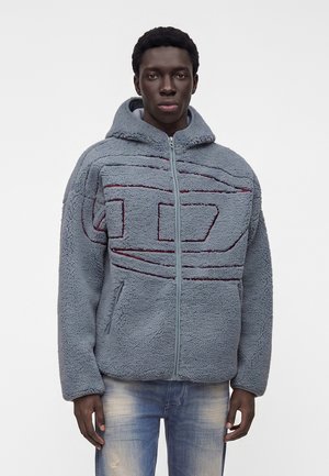 Jovem usando um casaco polar azul com capuz, com padrões geométricos vermelhos costurados e jeans azul claro desbotados, em pé contra um fundo branco.