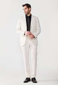 Weißer Anzug mit Schößchen-Revers, Ein-Knopf-Verschluss und Flat-Front-Hosen, kombiniert mit einem schwarzen Hemd und einer Krawatte sowie schwarzen Schuhen.