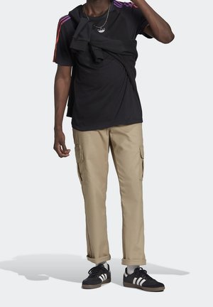 Man in een zwart Adidas T-shirt met meerkleurige strepen, beige cargobroek, zwart Adidas sneakers met witte strepen, en een zwart sweatshirt over de schouder.