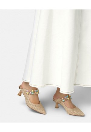Pies con zapatos mule beige tejidos con tacones y tiras adornadas con piedras de colores, combinados con una falda larga o vestido blanco.