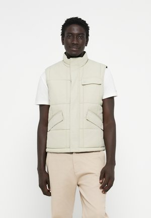 Gilet rembourré beige clair avec col haut, fermeture éclair à l'avant et deux poches latérales, porté par-dessus un t-shirt blanc et un pantalon beige.