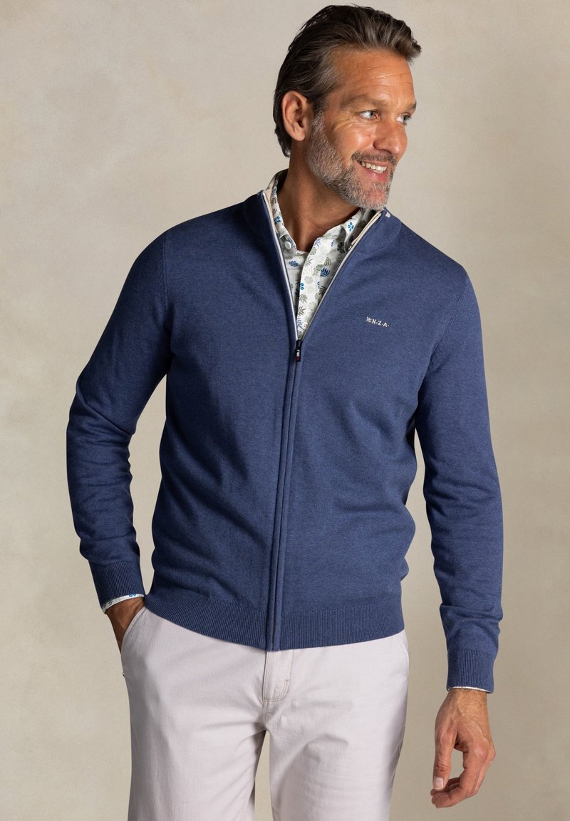 Blauwe gebreide zip-up sweater met een hoge kraag, geribde manchetten en onderrand. Draag het over een witte, bedrukte shirt, gestyled met lichtgekleurde broek.