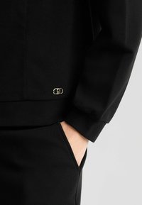 Zwarte sweatshirt met ribbelstructuur, voorzien van een klein metalen logo op de linkerzoom. Het ontwerp bevat een front pocket en aansluitende manchetten.