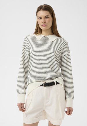 Femme portant un pull à rayures blanches et grises à manches longues avec un col blanc et un short blanc taille haute avec une ceinture noire.