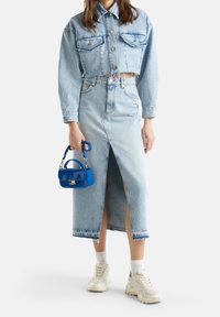 Krat i ljusblå denimjacka med två framfickor, i kombination med en matchande hög midjas kjol med slits framtill. Håller en liten blå handväska.