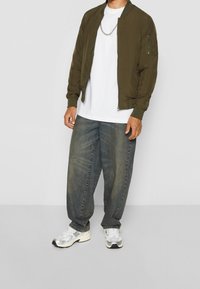 Olivgrön bomberjacka med ribbade manschetter, vit t-shirt, löst sittande blå denimjeans och silverfärgade träningsskor med svarta detaljer.