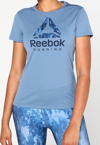 Maglietta sportiva azzurro chiaro con collo rotondo, caratterizzata da un logo Reebok blu scuro e un design astratto, realizzata in tessuto traspirante.