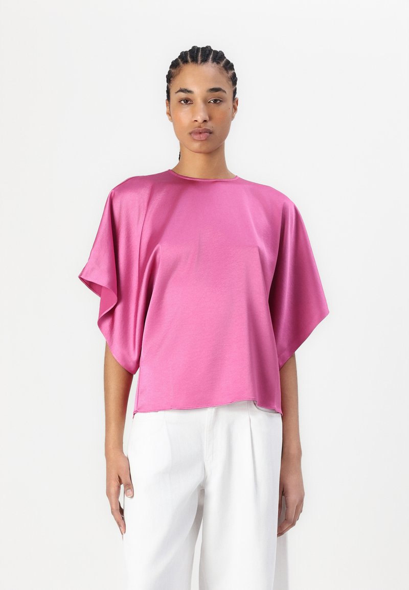 Fuchsia satintopp med løse batwing-ermer og rund hals, kombinert med hvite brede bukser. Glatt, skinnenede tekstur.