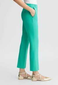 Pantaloni verdi su misura con tasche laterali, texture liscia e silhouette aderente. Indossati con scarpe a pianta dorate che mostrano un design a strap.