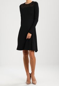 Vestito lungo nero a maniche lunghe con scollatura in pizzo e corpetto aderente. Orlo svasato e tessuto morbido, abbinato a tacchi alti beige.