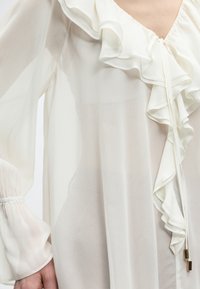 Διάφανη λευκή μπλούζα με βολάν στο V ντεκολτέ και μακριά μανίκια που συγκεντρώνονται στους καρπούς, φορεμένη στο πάνω μέρος του κορμού και το χέρι ενός ατόμου.