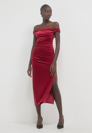 Frau in einem roten, schulterfreien Samtkleid mit einem Schlitz bis zum Oberschenkel, die schwarze High Heels trägt und vor einem weißen Hintergrund steht.