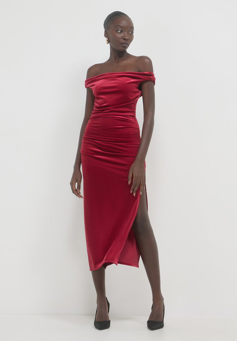 Frau in einem roten, schulterfreien Samtkleid mit einem Schlitz bis zum Oberschenkel, die schwarze High Heels trägt und vor einem weißen Hintergrund steht.