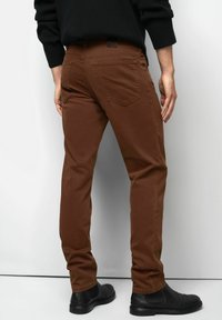 Man in een bruine slim-fit broek, zwart sweater en zwarte leren schoenen, staand met zijn rug naar de camera tegen een witte achtergrond.